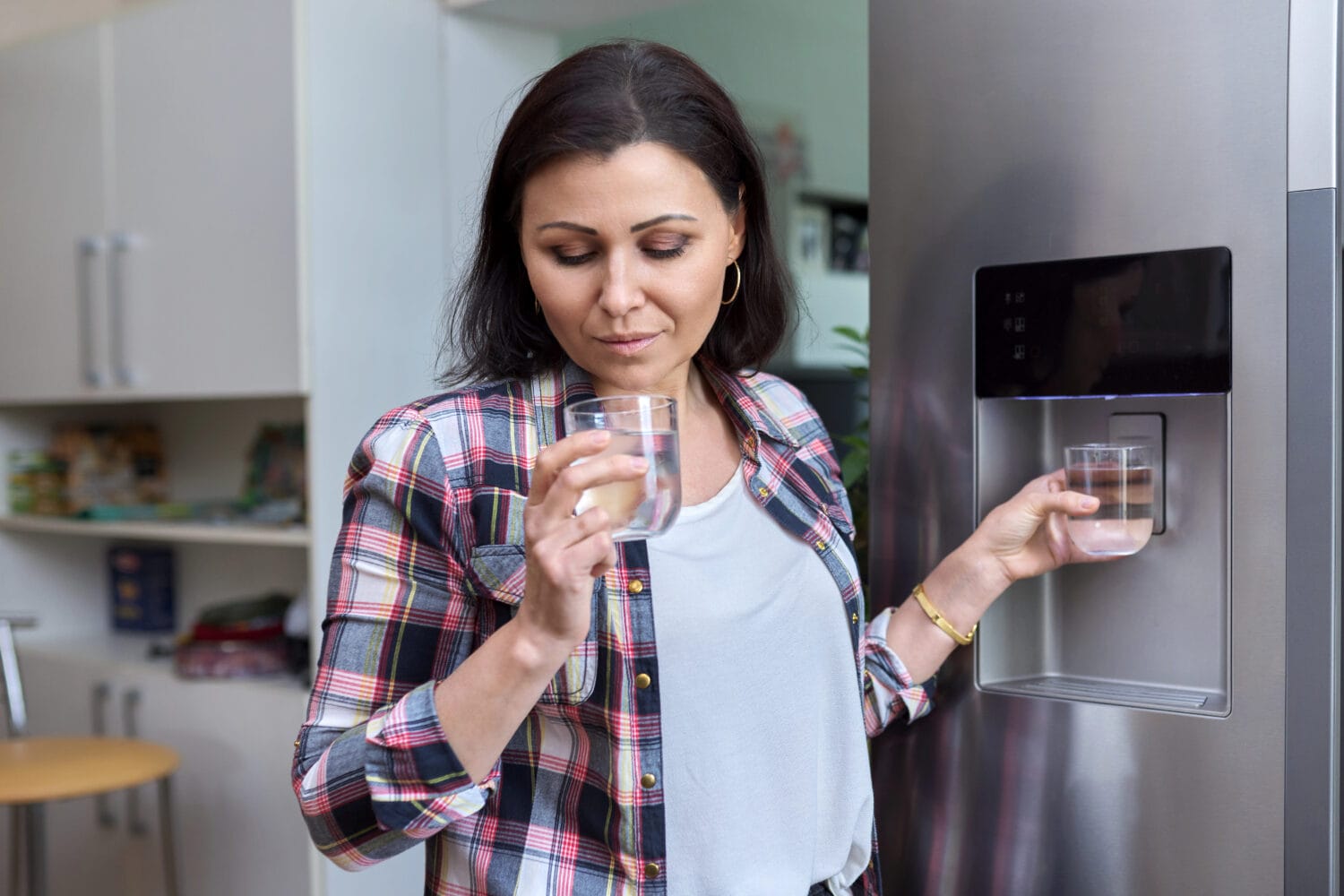 Woman standing by a stainless-steel refrigerator, holding a glass of water from the fridge dispenser and smelling it with a thoughtful expression.